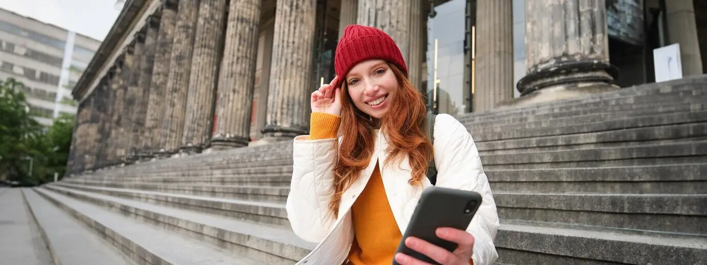 Retrato de una joven pelirroja con sombrero rojo sentada en unas escaleras; la turista mira su teléfono móvil mientras descansa.